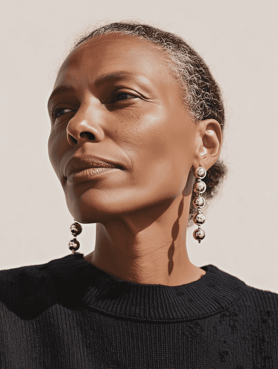 An elegant woman with a calm expression, wearing long, dangling earrings, looks into the distance. Her textured hair is pulled back, and she wears a black sweater.